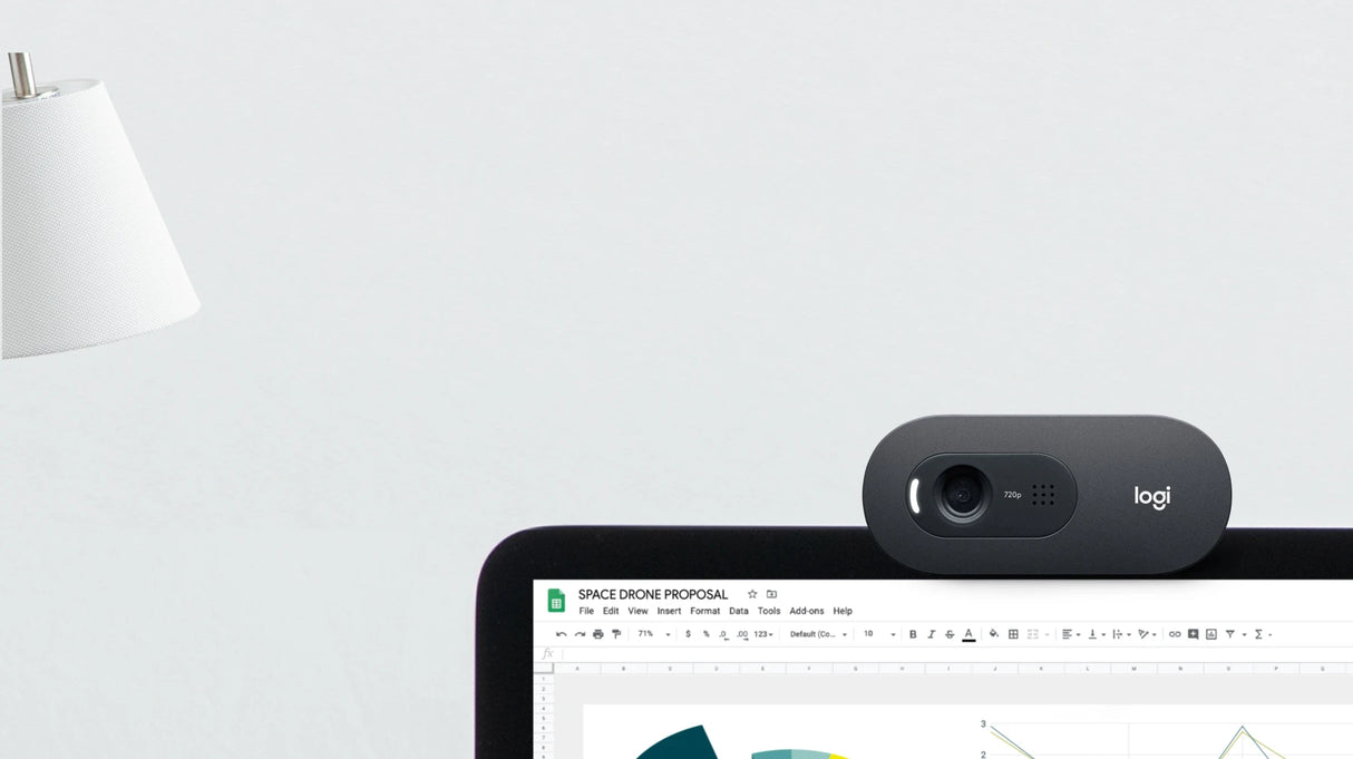 Lifestyle image of Logitech C505e HD webcam clipped to laptop screen during Google Meet video call, with status LED on, near a white lamp on light grey desk