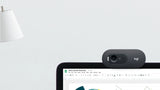 Lifestyle image of Logitech C505e HD webcam clipped to laptop screen during Google Meet video call, with status LED on, near a white lamp on light grey desk