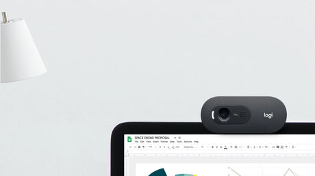 Lifestyle image of Logitech C505e HD webcam clipped to laptop screen during Google Meet video call, with status LED on, near a white lamp on light grey desk