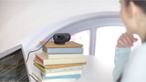 Logitech C505e HD webcam mounted on stack of books next to laptop on white desk, with person in blue shirt and arched window background, showing flexible setup versatility