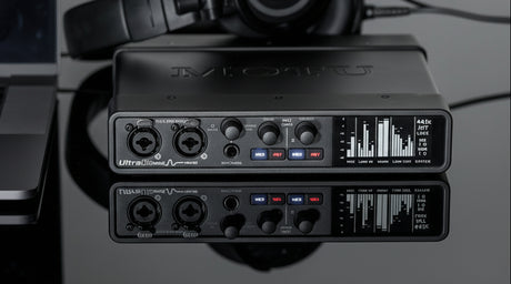 MOTU UltraLite mk5 black half-rack USB audio interface angled on glossy black surface next to a labtop, showing front XLR/TRS combo inputs with gain knobs, meters, headphone output, and illuminated buttons, with reflection below.
