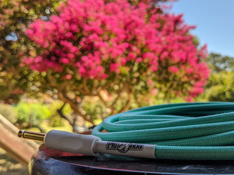 Pig Hog seafoam green woven instrument cable with white connector on wooden surface, pink flowering tree blurred in outdoor background