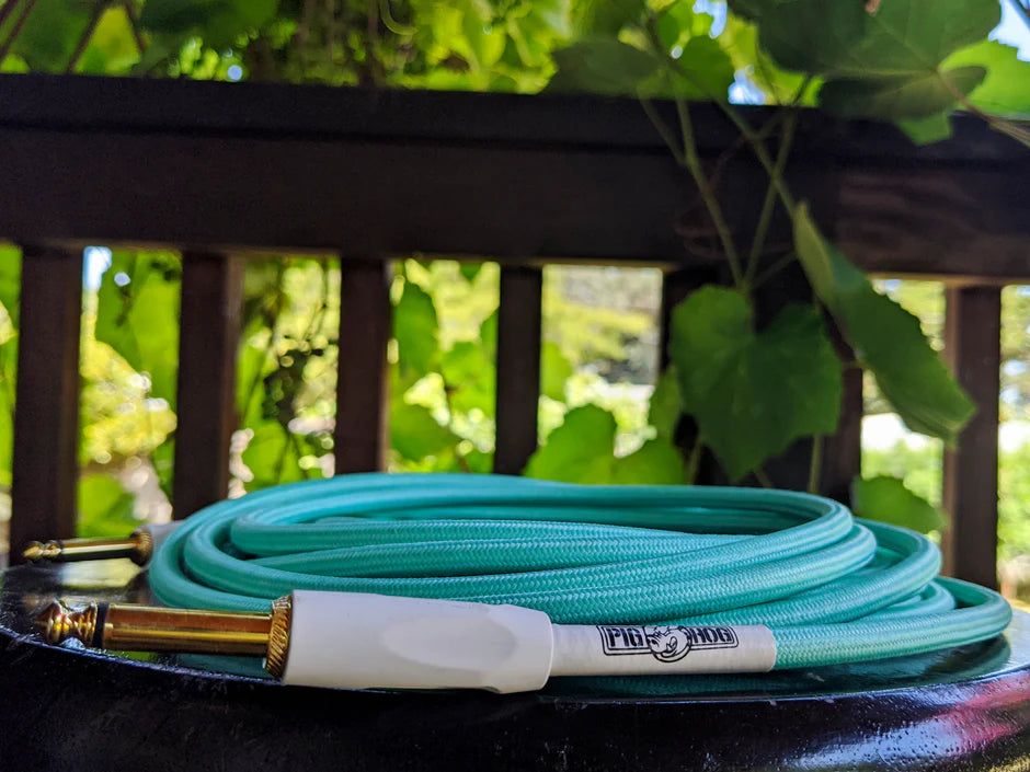 Pig Hog seafoam green woven instrument cable coiled on dark surface outdoors, green vines and railing blurred in background