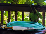 Pig Hog seafoam green woven instrument cable coiled on dark surface outdoors, green vines and railing blurred in background